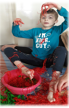 Chłopiec siedzi na dywanie przed sobą ma miskę z czerwonym i zielonym makaronem - bawi się
