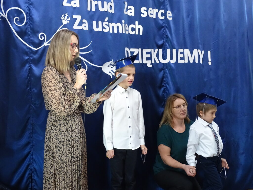 Nauczyciel mówi przez mikrofon obok niej stoi dwoje wychowanków mający birety na głowach