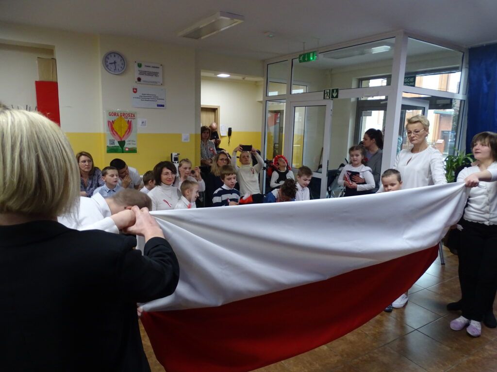 Na pierwszym planie znajduje się flaga Polski ułożona z długich pasów materiału białego                                 i czerwonego, którą trzymają dzieci w strojach galowych. W tle widać wychowanków i pracowników ośrodka.