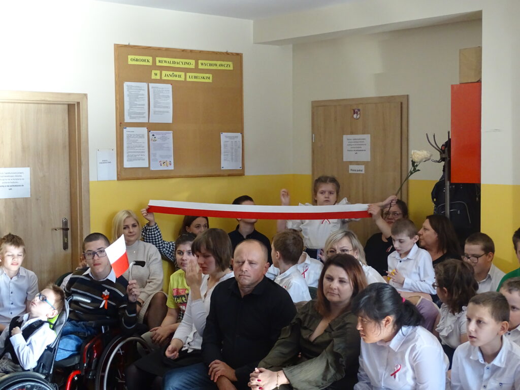 Cała społeczność Ośrodka jako widownia. Dzieci trzymają flagę Polski. Na pierwszym planie siedzi pani dyrektor ORW Iwona Miśkiewicz-Rachwał oraz pan dyrektor Poradni Psychologiczno-Pedagogicznej , Jarosław Lenart.