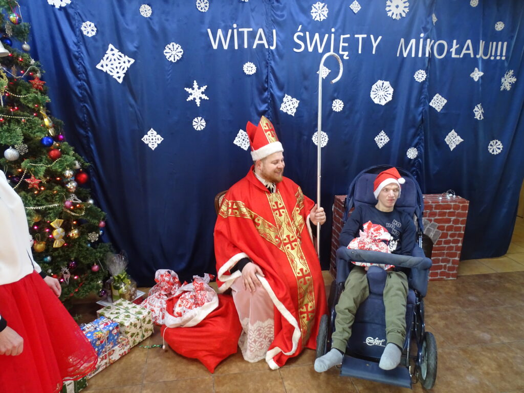 Mikołaj siedzi w fotelu, w ręku trzyma biskupią laskę. Po prawej stronie znajduje się chłopak na wózku z prezentem na kolanach. Po lewej stronie na podłodze widać worek  Mikołaja i prezenty pod choinką.  W tle widnieje biały napis WITAJ ŚWIĘTY MIKOŁAJU !!! na granatowym materiale, wokół niego białe śnieżynki.
